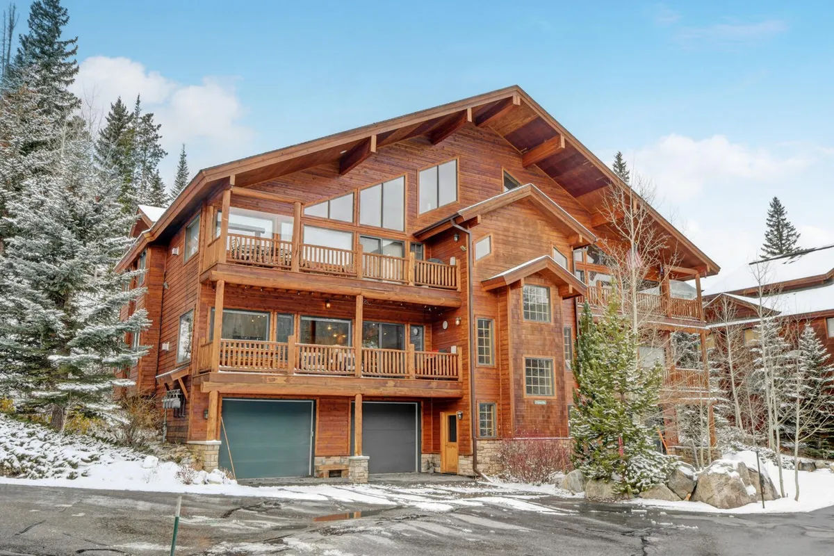 The image showcases the inviting wooden exterior of a luxury condo nestled among snow-dusted trees. Large windows and spacious balconies overlook the scenic Winter Park landscape, hinting at cozy evenings indoors and panoramic mountain views. The overall mood is warm and welcoming, inviting guests to picture themselves gathering after a day on the slopes.