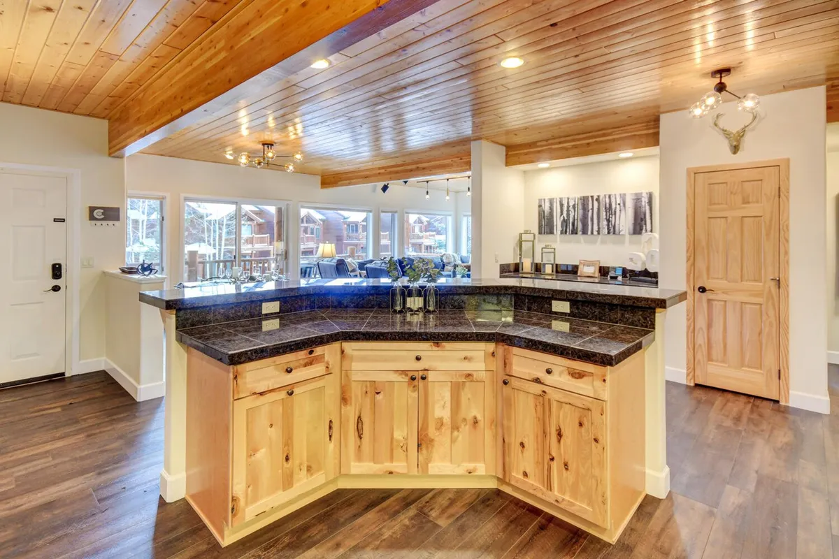A modern kitchen island with a granite countertop, wooden ceiling with custom paneling, and large windows bringing in natural light. Bar stools invite guests to gather, and the space feels both luxurious and practical for entertaining or crafting celebratory drinks.
