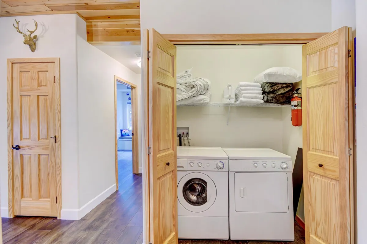 A functional and welcoming laundry area with a washing machine, dryer, and shelves stocked with towels. Warm wooden door frames and organized storage convey a sense of homey convenience, ideal for longer stays.