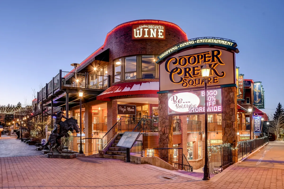 The lively exterior of Cooper Creek Square, highlighted by glowing decorative lights, visible restaurant and wine shop signs, and an inviting outdoor seating area. The atmosphere is festive and social, perfect for dining, shopping, and enjoying community events in Winter Park town.