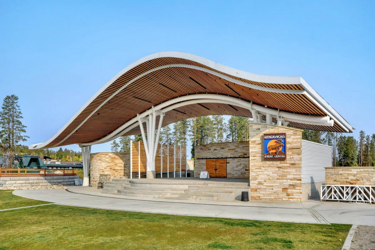 An exterior view of a striking event center featuring a curved roof structure, prominent stone wall, wooden beams, and a welcoming event center sign. The outdoor space is set up for gatherings, concerts, and community events, exuding a festive and lively mood.