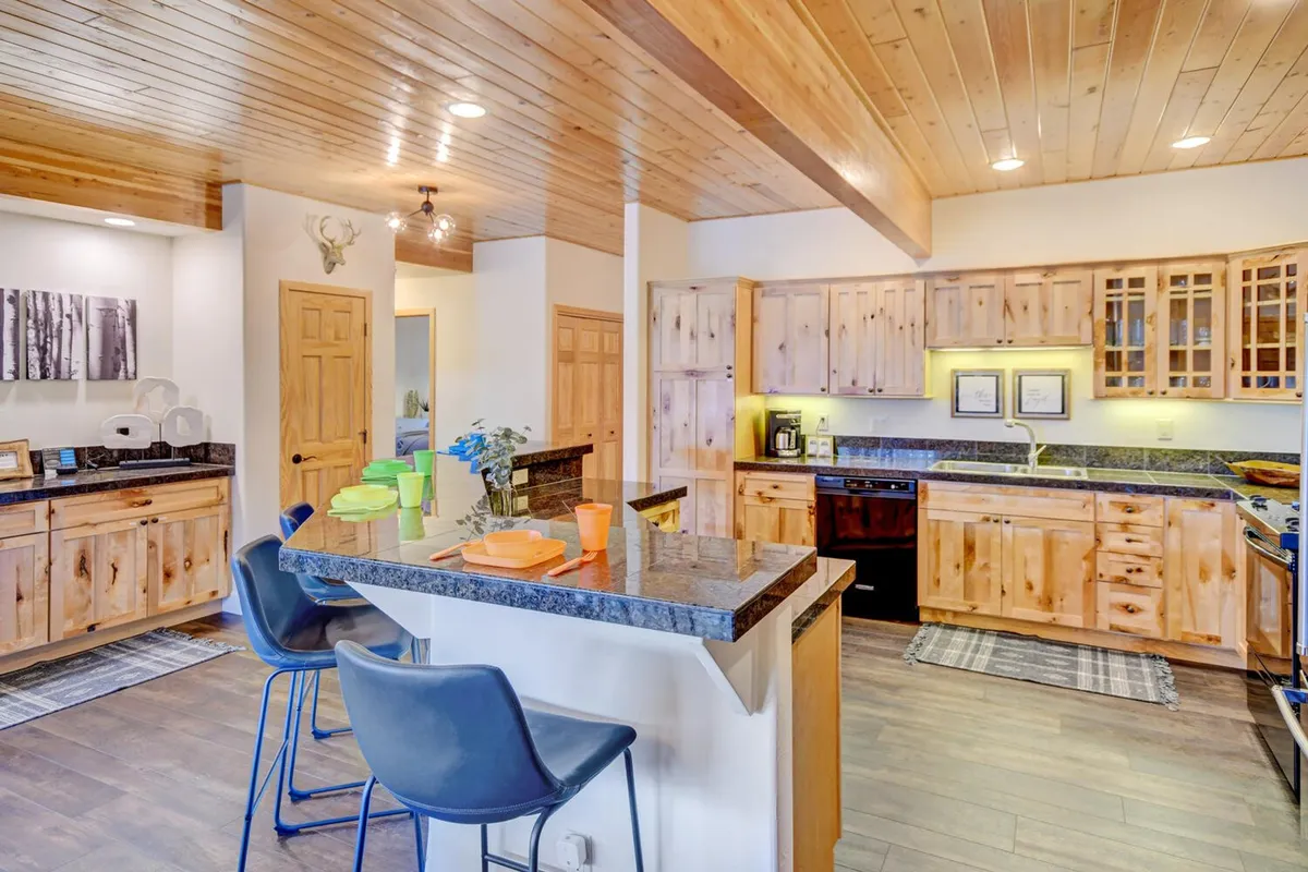 A spacious, well-lit kitchen featuring a long granite countertop, modern wooden cabinets, bar stools lined up at the island, visible kitchen appliances, and thoughtful decorative artwork. The open layout provides ample space for cooking and entertaining, with a friendly, inviting atmosphere perfect for group gatherings.