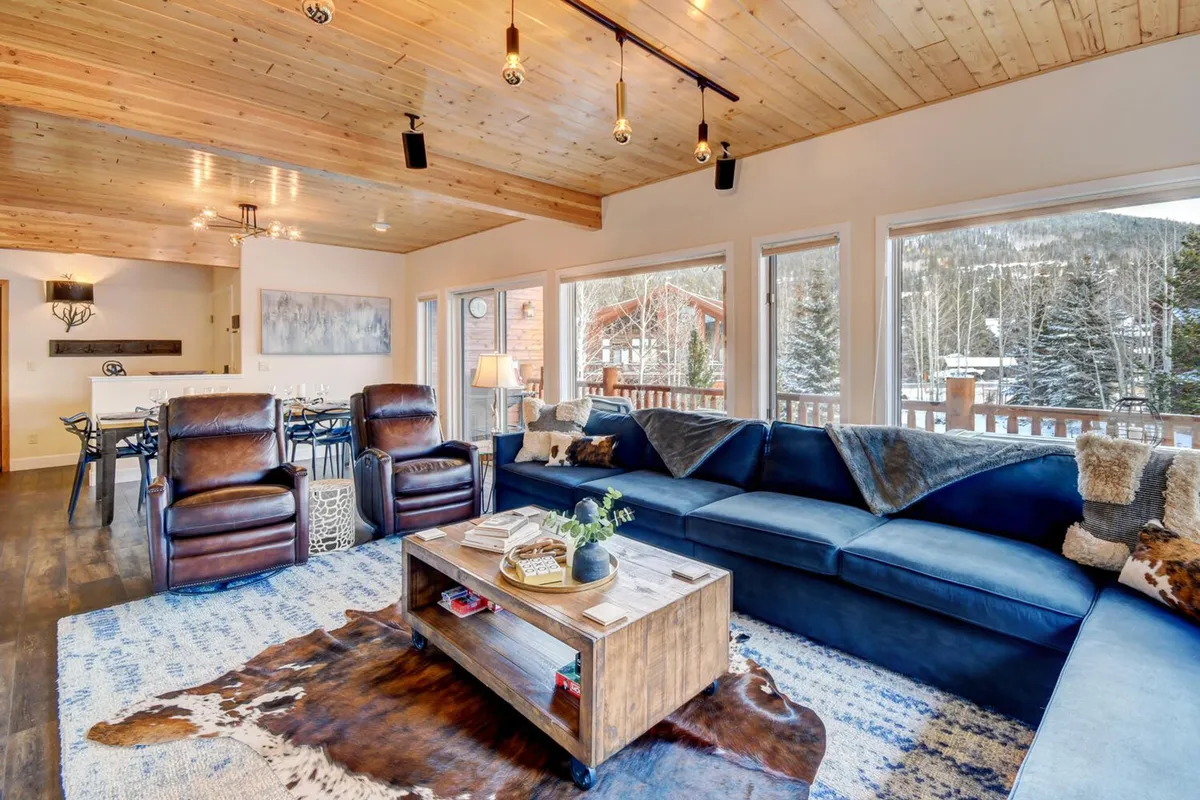 A spacious, inviting living room with a large blue sofa, wooden coffee table, and comfy leather chairs arranged for conversation. The setting features large windows with mountain views, allowing natural light to fill the space. The arrangement is perfect for relaxing with family, playing board games, or enjoying cozy evenings together.