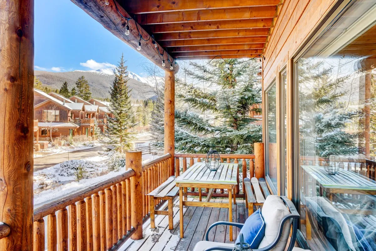 A wooden deck furnished with a table and chairs, surrounded by mature trees and offering captivating mountain views. The deck is spacious, cozy, and set up for relaxing, dining, or entertaining outdoors in any season.