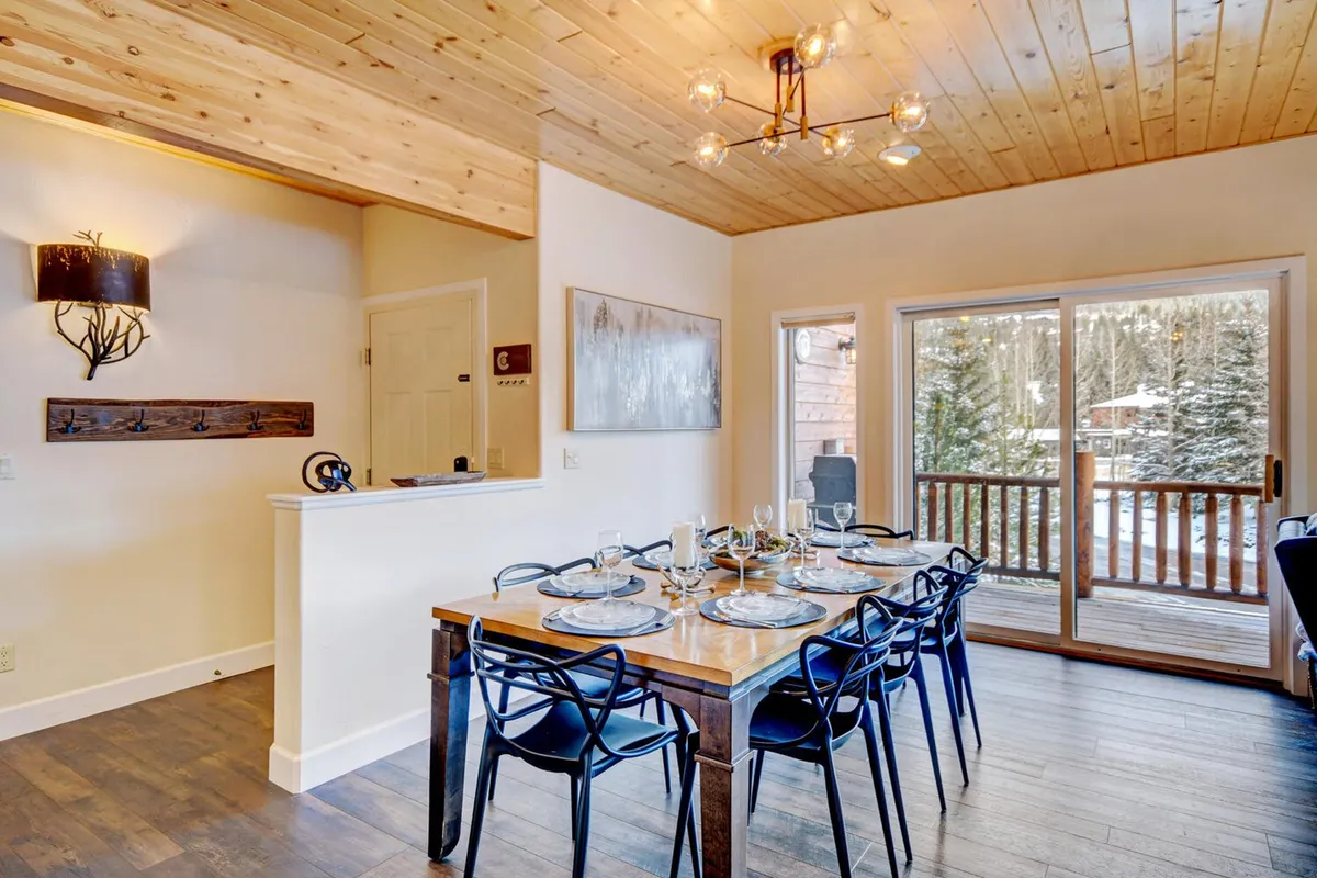 A bright dining area with a rustic wooden table set beneath an elegant chandelier, framed by large windows that let in abundant natural light. The wooden ceiling and spacious layout create a warm, communal environment ideal for shared meals and celebrations.