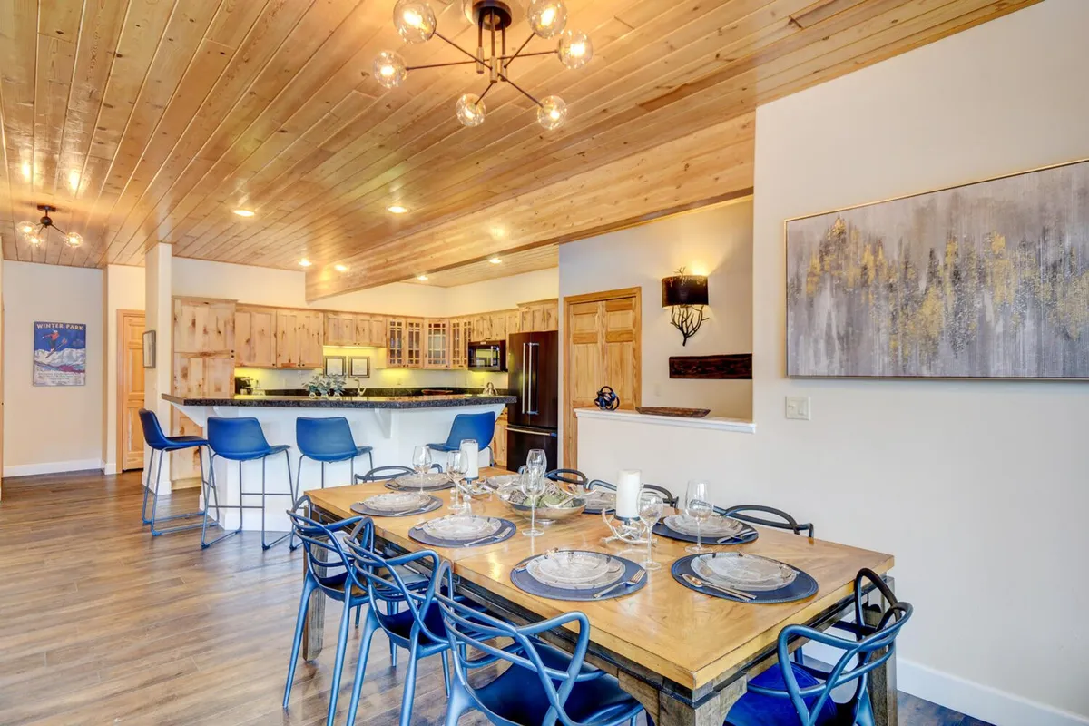 A well-lit dining area showcasing a dining table that seats eight, a stylish chandelier above, and a kitchen island with wooden cabinetry and bar stools nearby. The layout seamlessly connects the spaces, ideal for group meals or casual snacks after a day on the slopes.