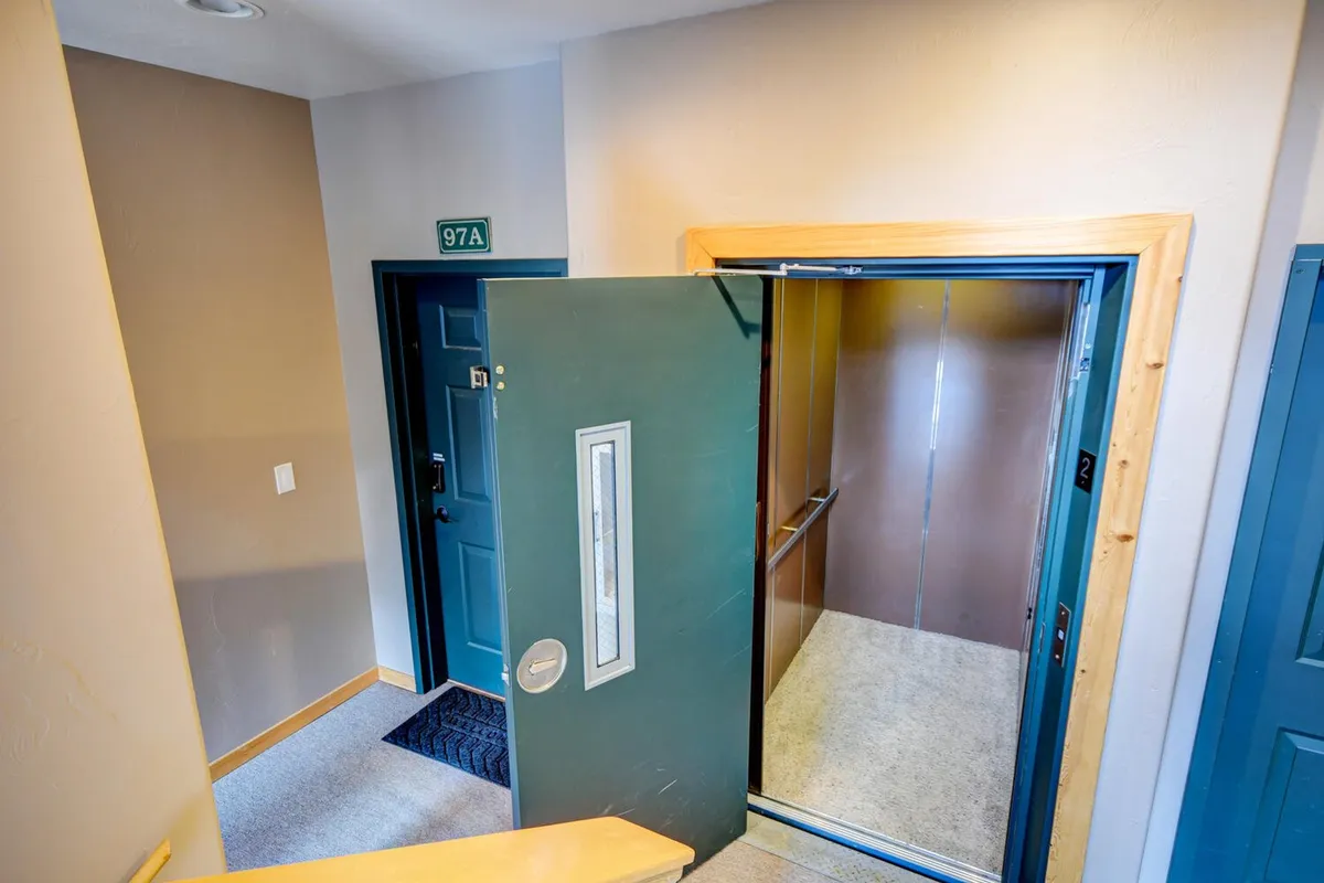 This image shows a green door at the elevator entrance, with the unit number '97A' visible. The scene suggests the excitement of arriving or departing, and hints at the building’s connection to broader adventures outside the condo.