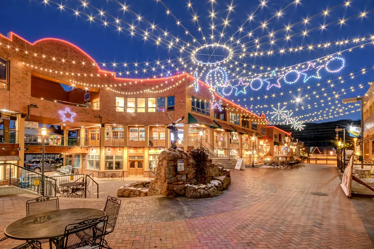 An animated village square with decorative lights, restaurant signage, outdoor seating, and a bubbling fountain. The area buzzes with energy from socializing, festive celebrations, and community events.